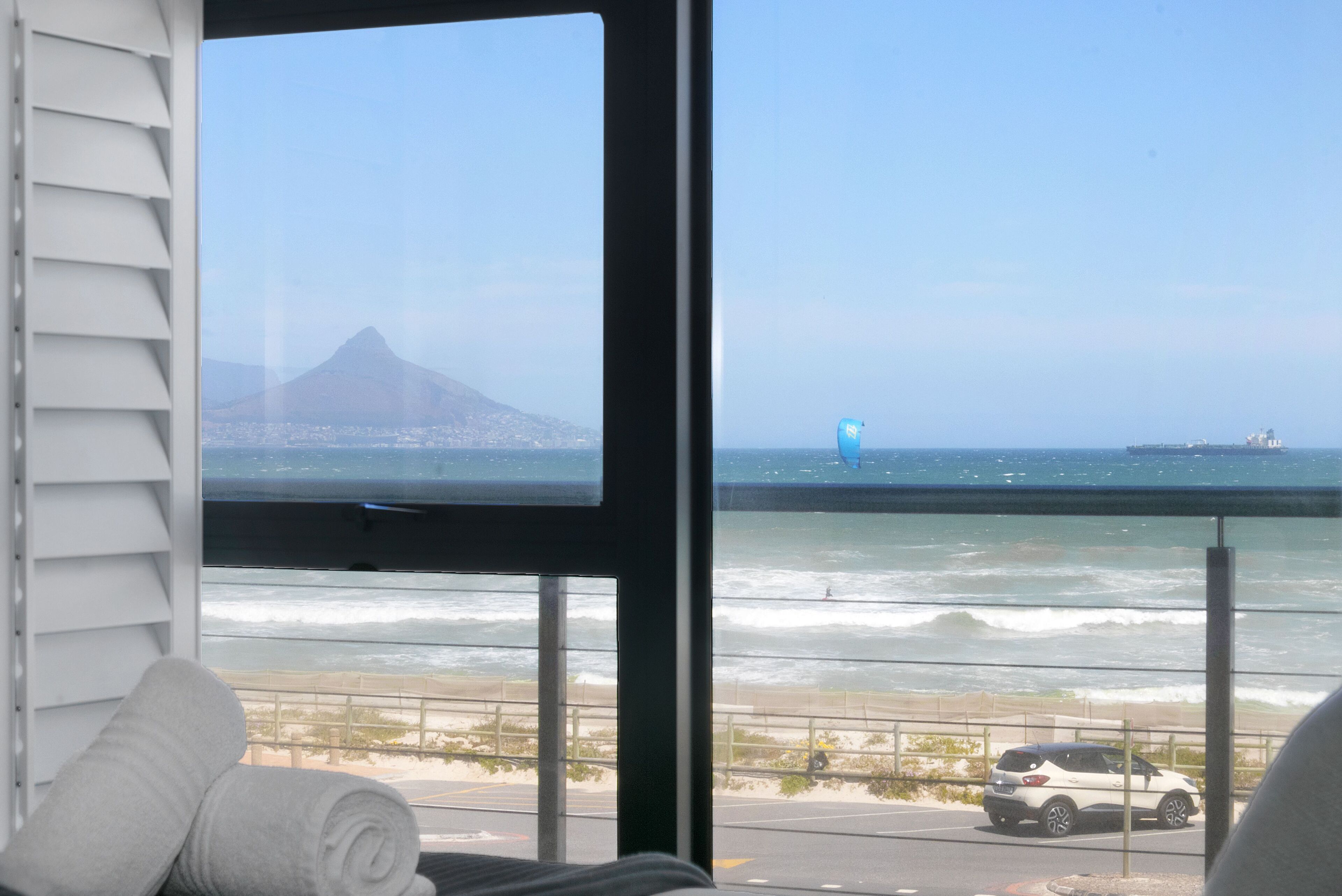 A room with a large window overlooking a beach and ocean, a white car parked outside, and rolled towels on a bed.