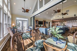 A spacious living area with a large dining table, wicker chairs, a kitchen in the background, and ceiling fans.