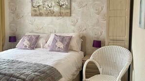 A bedroom with a bed, two pillows, a fluffy blanket, a wicker chair, a nightstand, a lamp, and a floral wallpaper.