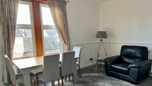 A room with a dining table and chairs, a black leather sofa, a floor lamp, and a view through a window with curtains. - Charming Seaside home. sleeps 4 with free parking (Dundee)