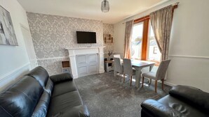 A living room with a fireplace, a TV mounted above it, a dining area with a table and chairs, and a sofa. - Charming Seaside home. sleeps 4 with free parking (Dundee)
