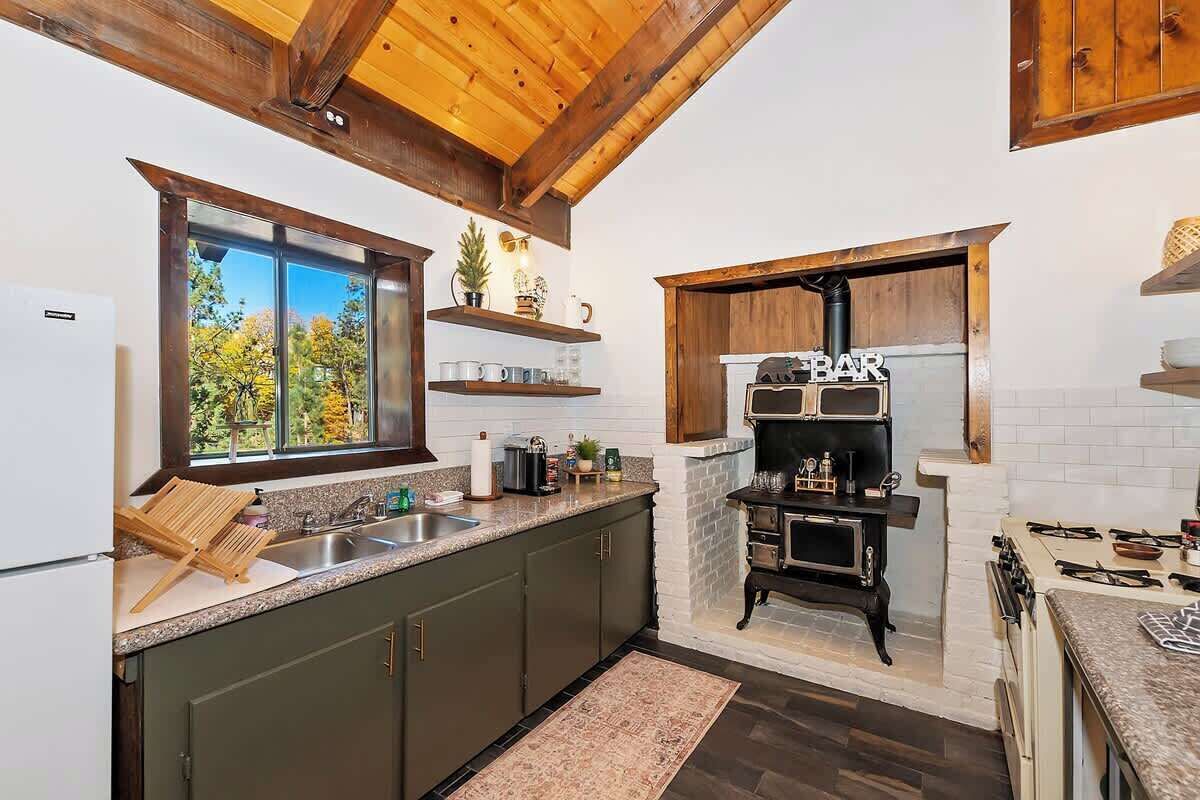 Una cocina con armarios verde oscuro, techo de madera, ventana con vista a árboles, estufa empotrada y una repisa con tazas.