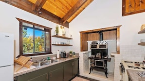 A kitchen with dark green cabinets, a wooden ceiling, a window with a view of trees, a built-in stove, and a shelf with mugs. - Closer to the Stars Fire Pit Dog-Friendly (Big Bear Lake)