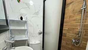 Un baño con lavamanos, inodoro y ducha blancos. Hay un estante pequeño con una planta y una botella de jabón. El suelo está cubierto de baldosas, y hay una alfombra marrón.