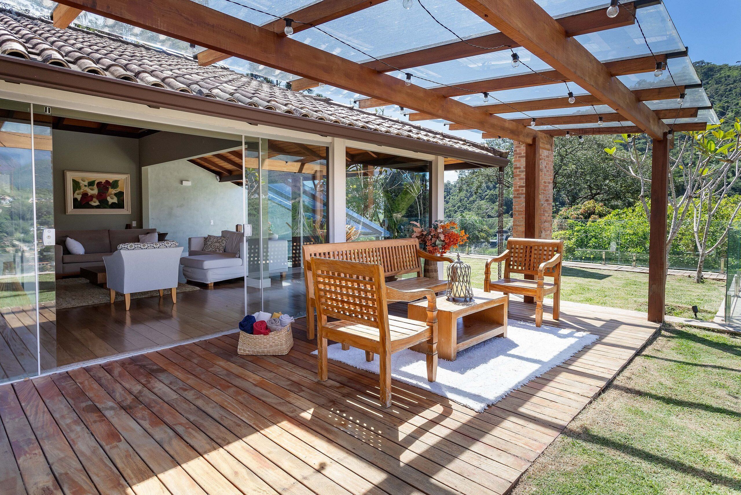 A covered patio with wooden furniture, a glass wall, and a view of a garden.