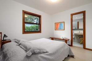 A bedroom with a large bed, a bedside table with a lamp, a small bench, and a window with a view of greenery. - Magnificent views, 3 suites, pool w/ heated tub (Petrópolis)