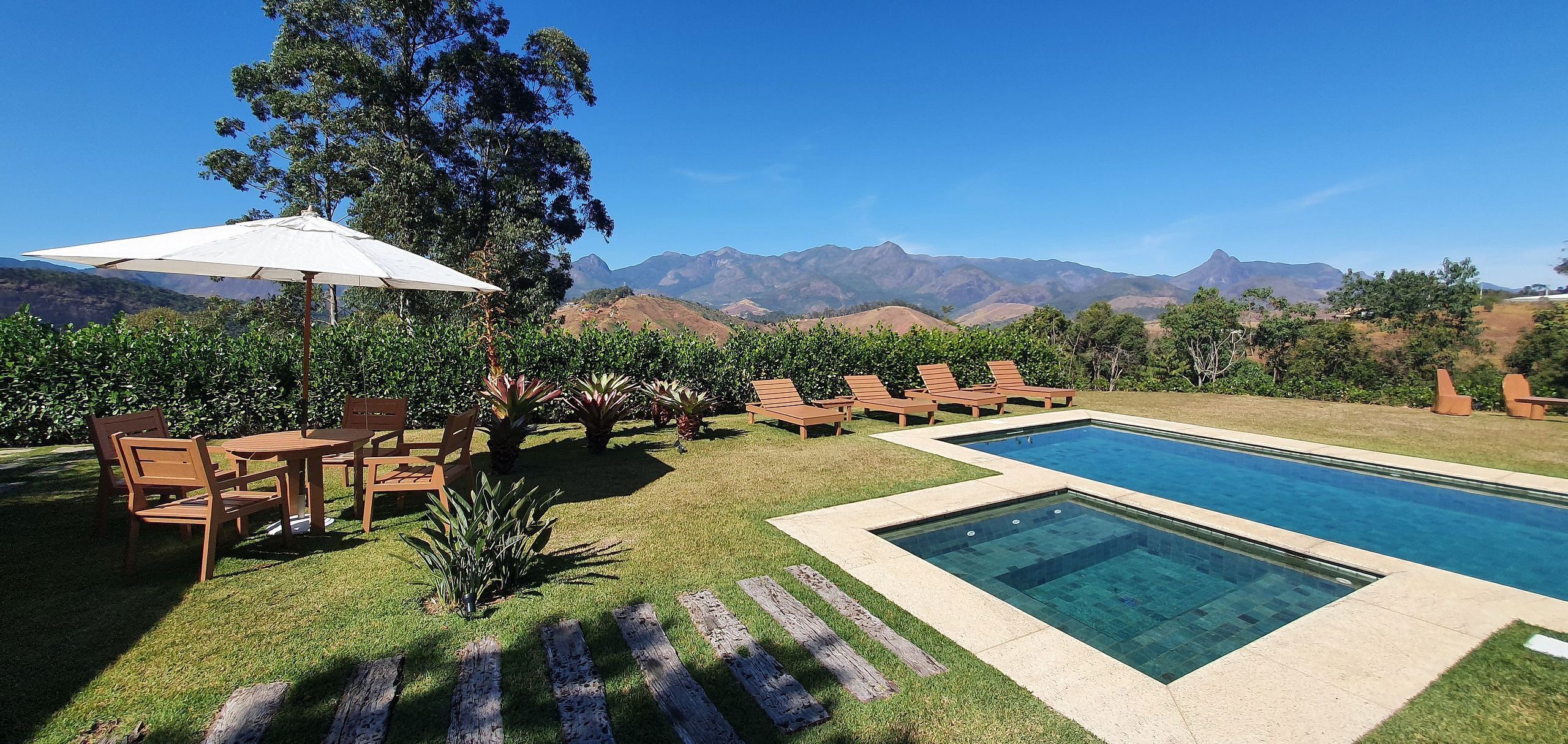 Un espace piscine doté de chaises longues et d’un parasol, entouré de verdure et avec des montagnes en toile de fond.