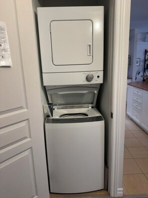 A compact, white laundry machine in a narrow space, with a door on the left and a kitchen area visible in the background. - Lake Archambault, Apartment 316 (Saint-Donat-de-Montcalm)