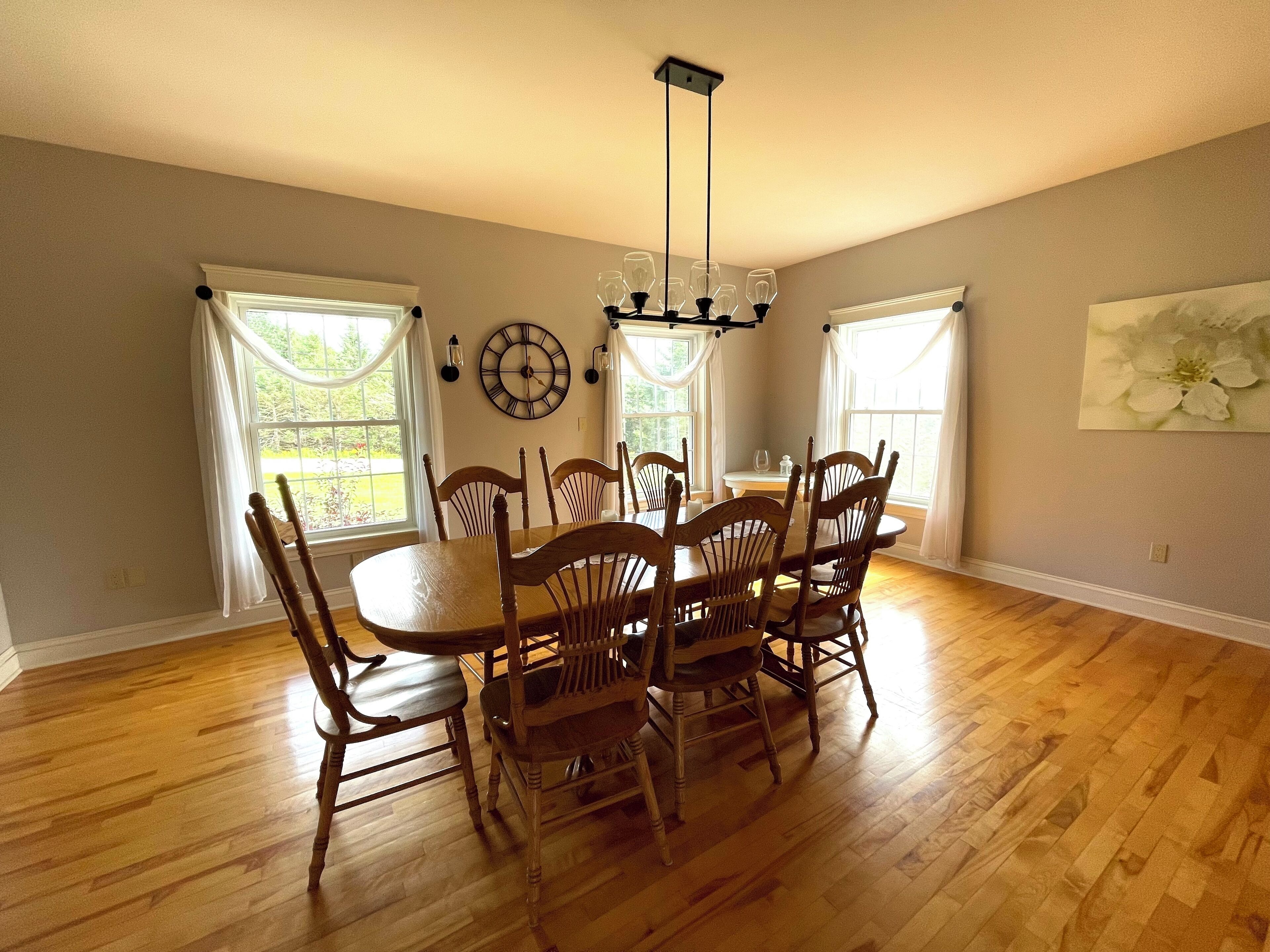 A dining room with a wooden table and chairs, large windows with white curtains, a wall clock, and a hanging light fixture.