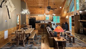 A rustic cabin interior with a wooden ceiling, a stone fireplace, a dining area with a wooden table and benches, a sofa, and a kitchen with wooden cabinets and a stone accent wall. - River Shoals Cabin! Hot Tub-Water front (Cleveland)