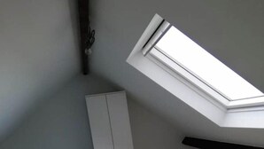 Ett sovrum med ett sluttande tak, ett takfönster, en säng med ett blått, blommigt sängöverkast, en garderob och en bänk.