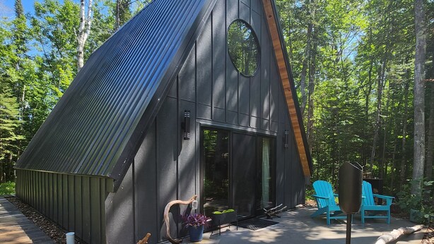 Eine moderne Hütte mit geneigtem Dach, großen Fenstern und einer Terrassenfläche mit Sitzgelegenheiten und Topfpflanzen.