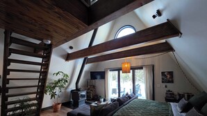 Ein Schlafzimmer im Loft-Stil mit Schräge, einer Holztreppe, einem großen Bett mit gemustertem Überwurf, einer Topfpflanze und Blick ins Freie durch ein Fenster.