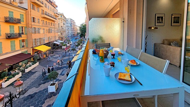 Varanda com mesa posta para o café da manhã, vista para uma rua com edifícios e lojas.