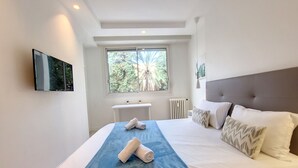 Quarto de hotel moderno com uma cama grande, televisão na parede e janela com vista para árvores.
