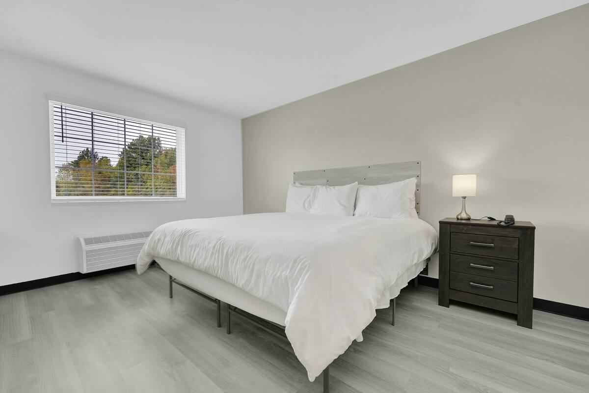 A bedroom with a bed, a bedside table, a window with blinds, and a radiator.