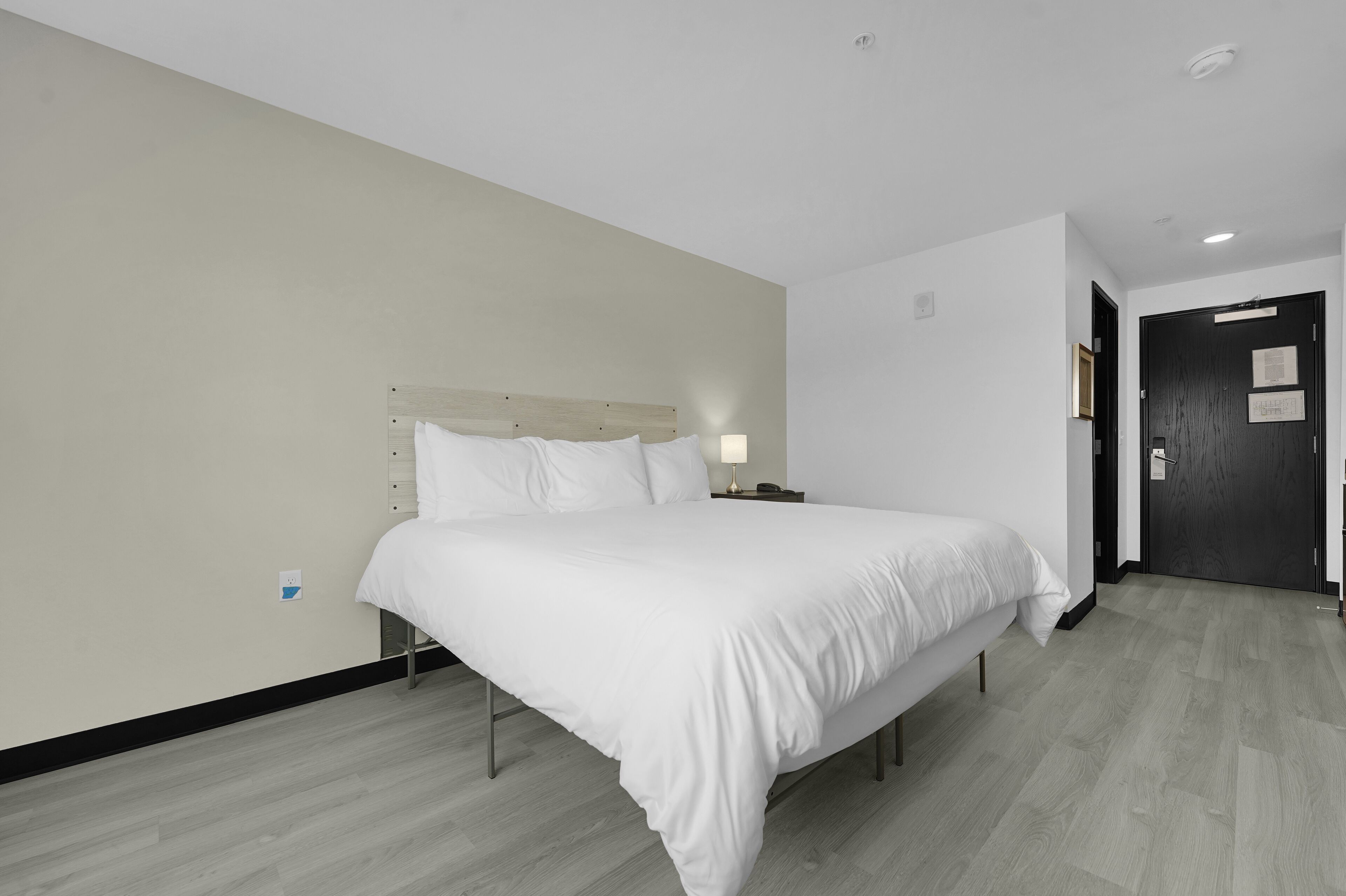 A modern hotel room with a large bed, a bedside table with a lamp, and a dark door.