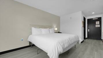 A modern hotel room with a large bed, a bedside table with a lamp, and a dark door.