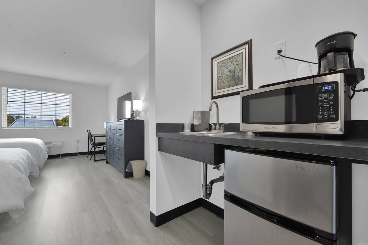 A modern hotel room with a kitchenette, a bed, a desk, and a window with a view of the outside.
