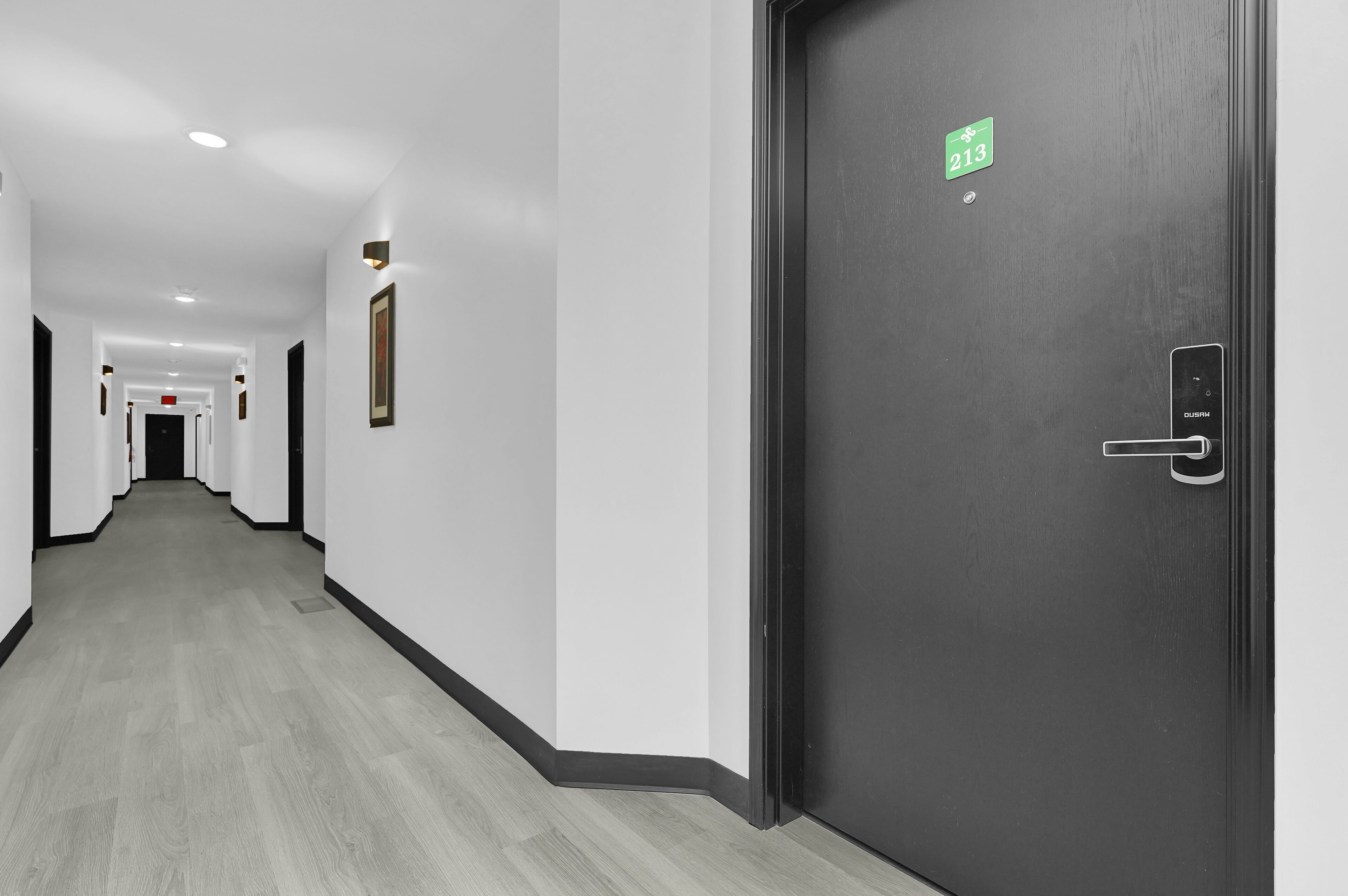 Un pasillo de hotel con una puerta oscura marcada con el número 213, un suelo de madera claro y varias fotografías enmarcadas en las paredes.