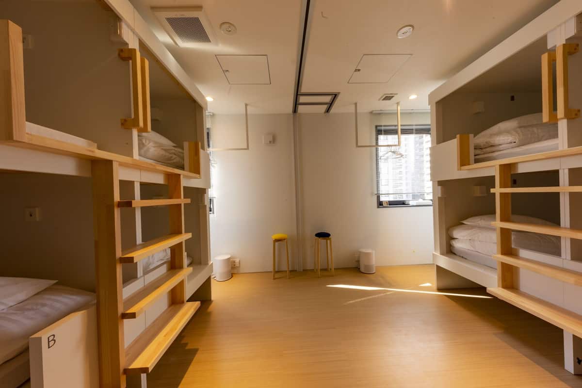 Quarto em dormitório com beliches, um banquinho, uma lixeira e uma janela com persianas.