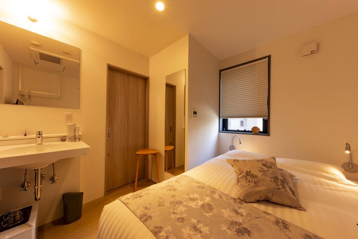 Quarto de hotel com cama, mesa de cabeceira, espelho, janela com persianas e banheiro com pia e espelho.