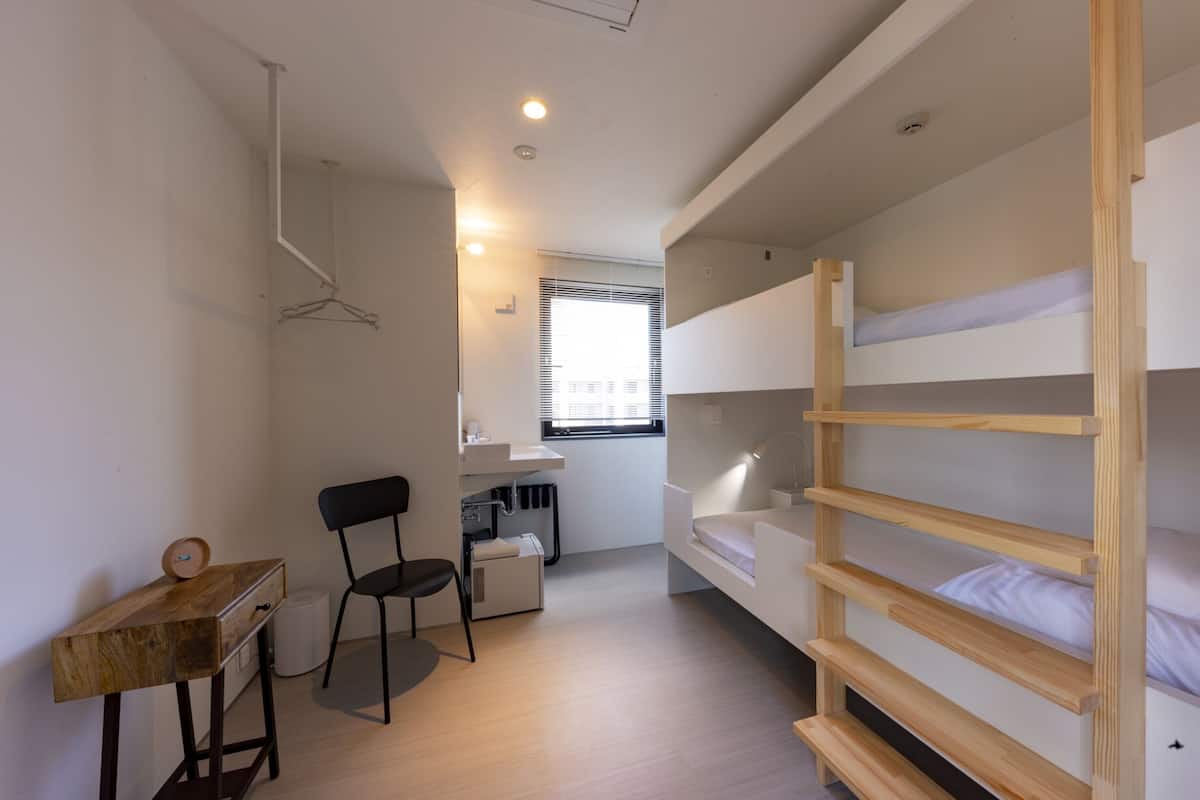 Quarto compacto com beliches, escrivaninha, cadeira e janela com persianas.