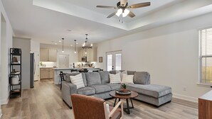 A spacious living room with a grey sectional sofa, a brown leather chair, and a coffee table. A ceiling fan and pendant lights are visible. - Newly built home minutes from Texas A&M campus (College Station)