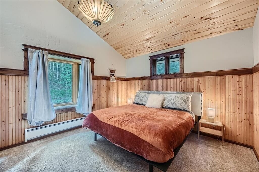 A bedroom with a wooden ceiling, a large bed with a red blanket, a nightstand, and a window with curtains.