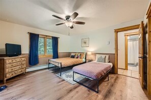 Une chambre avec deux lits, un ventilateur de plafond, une télévision, une commode et une fenêtre avec des rideaux.