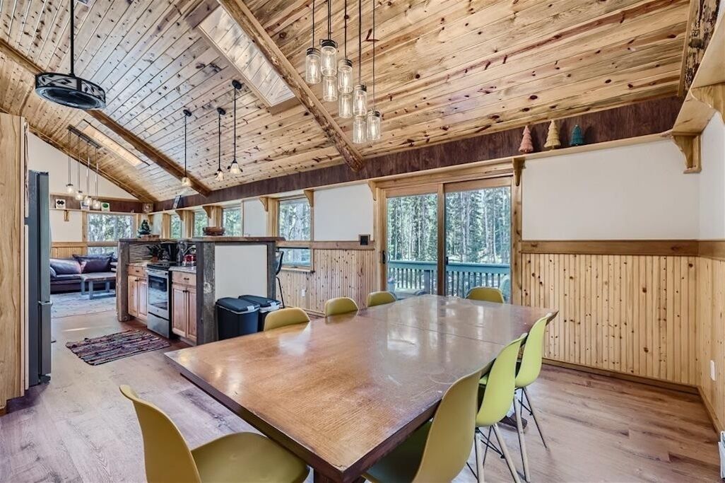 A spacious dining area with a wooden table and chairs, a kitchen in the background, and large windows with a view of trees.
