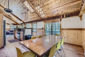 Un grand espace à manger avec une table et des chaises en bois, une cuisine à l’arrière-plan et de grandes fenêtres donnant sur la forêt.
