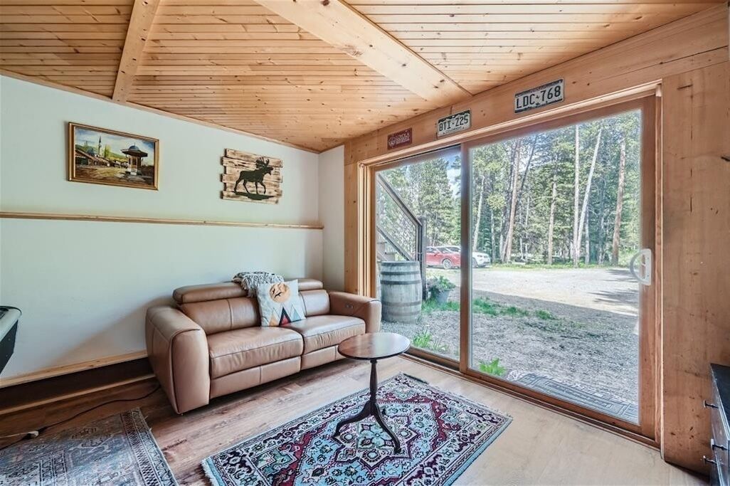 A wooden cabin interior with a beige leather sofa, a patterned rug, and a large sliding glass door leading to an outdoor area with trees and a red vehicle.