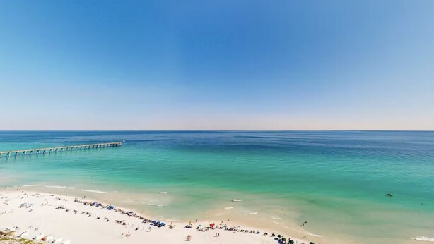 A beach with a pier extending into the water, a sandy shore with umbrellas and people, and clear blue skies.