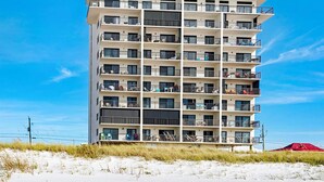 A multi-story hotel with balconies, located near a beach with sand dunes.