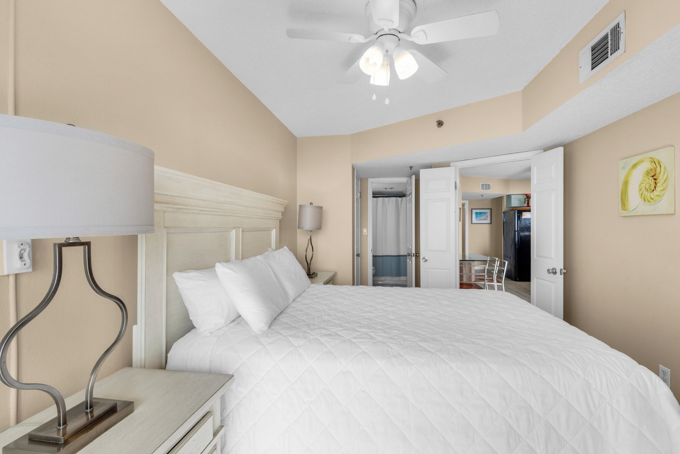 Une chambre d’hôtel comprenant un lit, une table de chevet, une lampe, un ventilateur de plafond et une porte ouverte donnant sur une salle de bain et une kitchenette.