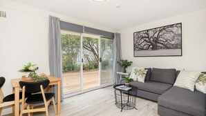 Un salon moderne avec un canapé gris, une table à manger avec des chaises, une petite table d’appoint et une grande fenêtre donnant sur des arbres.