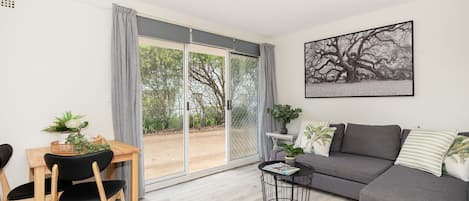 A modern living room with a grey sofa, a dining table with chairs, a small side table, and a large window with a view of trees.