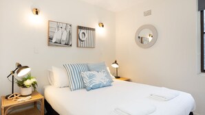 Une chambre à coucher bien rangée, avec un lit, deux lampes de chevet, une petite table avec une lampe et une plante, et deux œuvres d’art accrochées au mur.