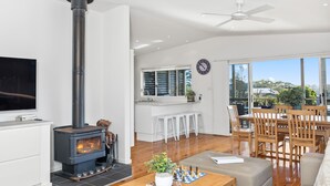 A modern living room with a fireplace, a dining area with wooden furniture, and a kitchen with a large window.