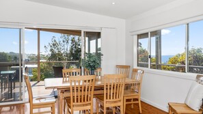 Un coin repas avec une table et des chaises en bois, de grandes fenêtres donnant sur les arbres et la mer, et un balcon avec une balustrade.
