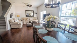 A spacious living room with a dark wood dining table, beige sofas, and a large window with blinds.