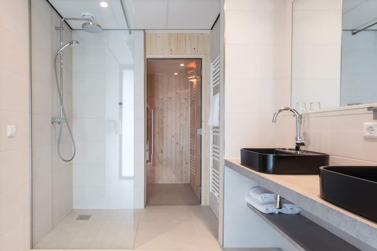 Un bagno moderno con doccia vetrata, una porta in legno che dà sulla sauna e un lavandino nero con specchio.