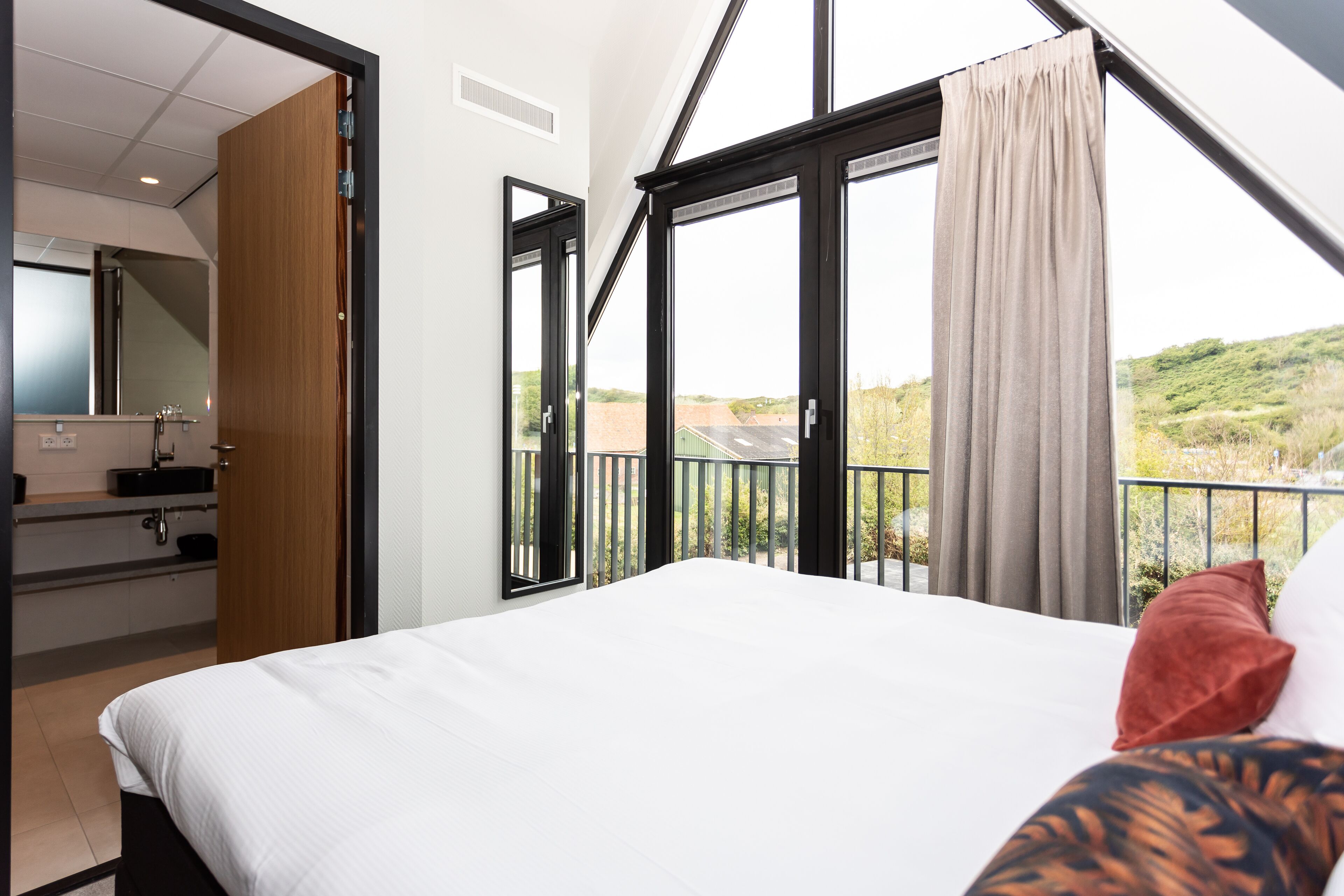 A bedroom with a large bed, a view of a balcony, and a bathroom with a mirror and sink.