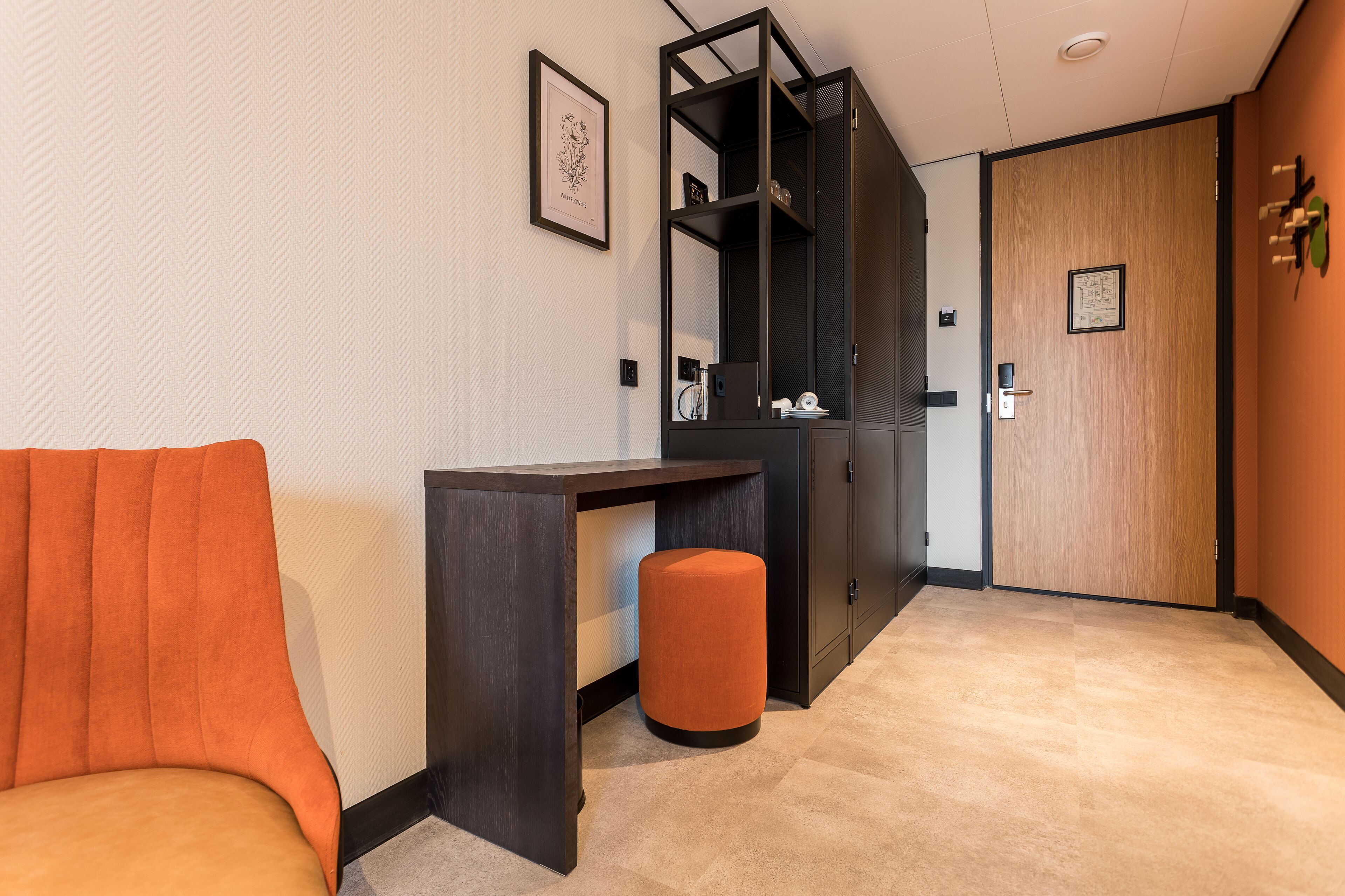 A modern hotel room with a sofa, a desk, a small stool, and a wardrobe.
