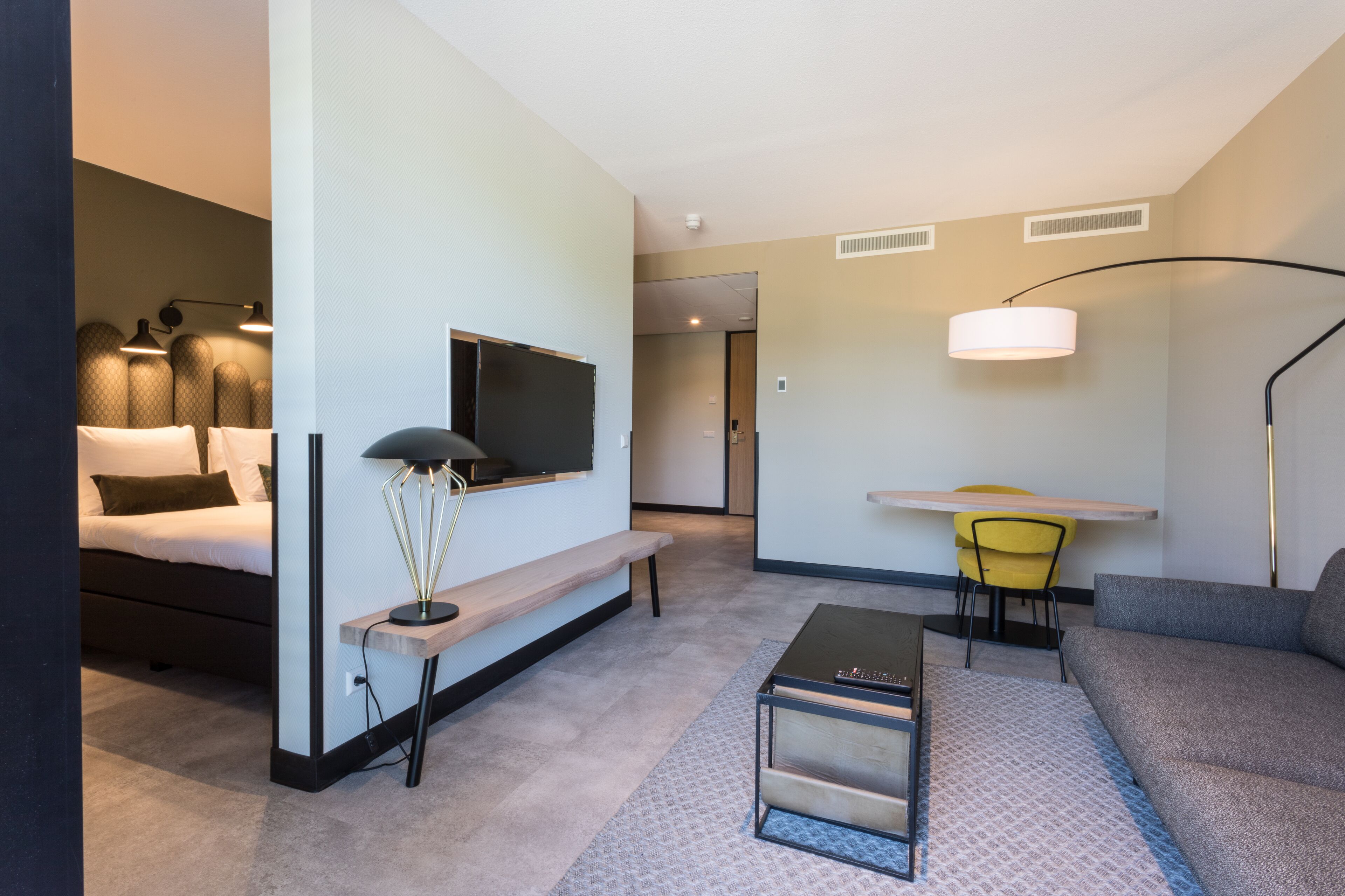 A modern hotel room with a bed, a desk, a sofa, a lamp, and a TV.