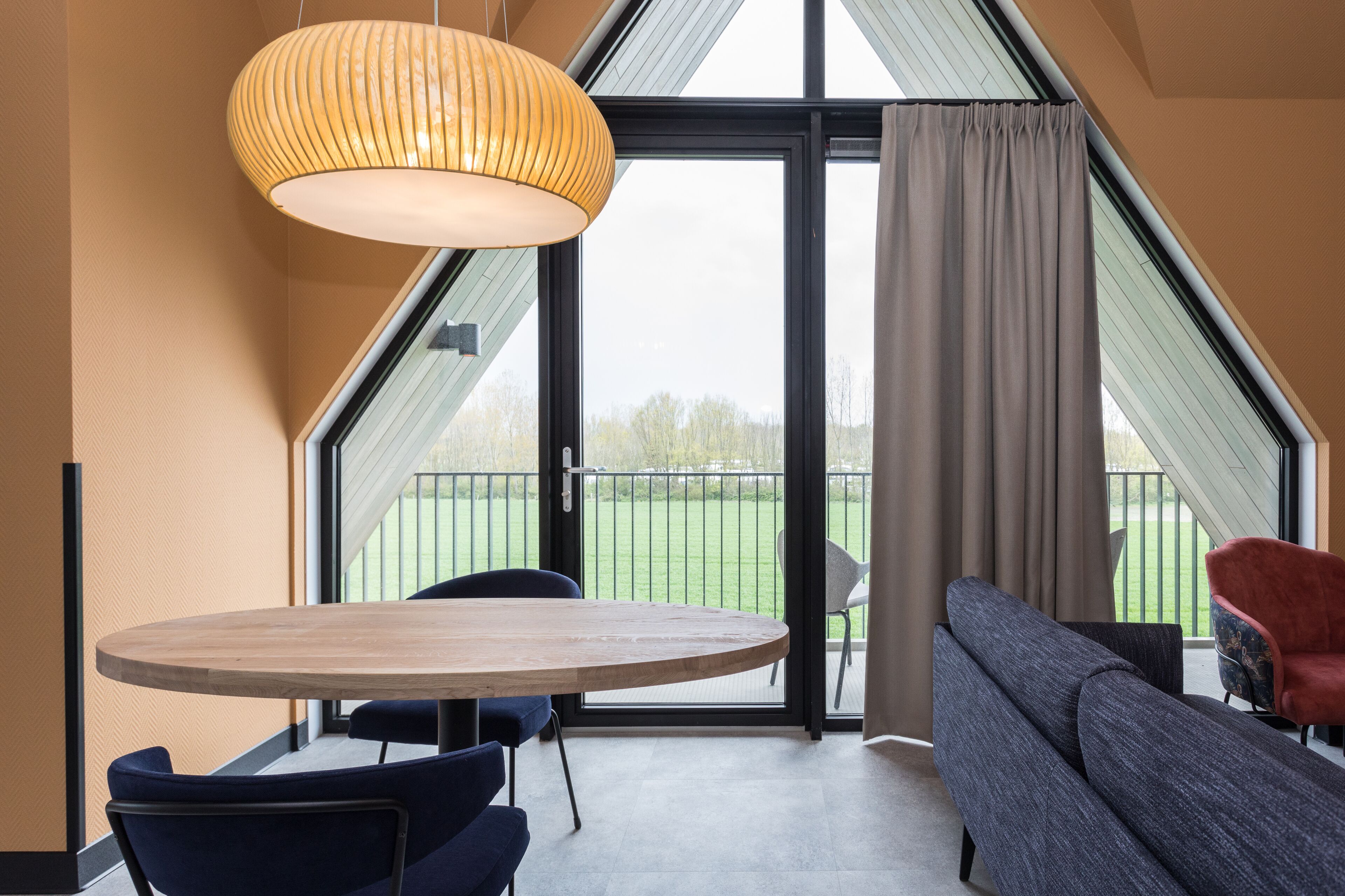A modern room with a round wooden table, two chairs, a sofa, and a large window with a view of a green field.