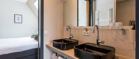 A modern hotel room with a large bed, a double sink, a mirror, and a window.