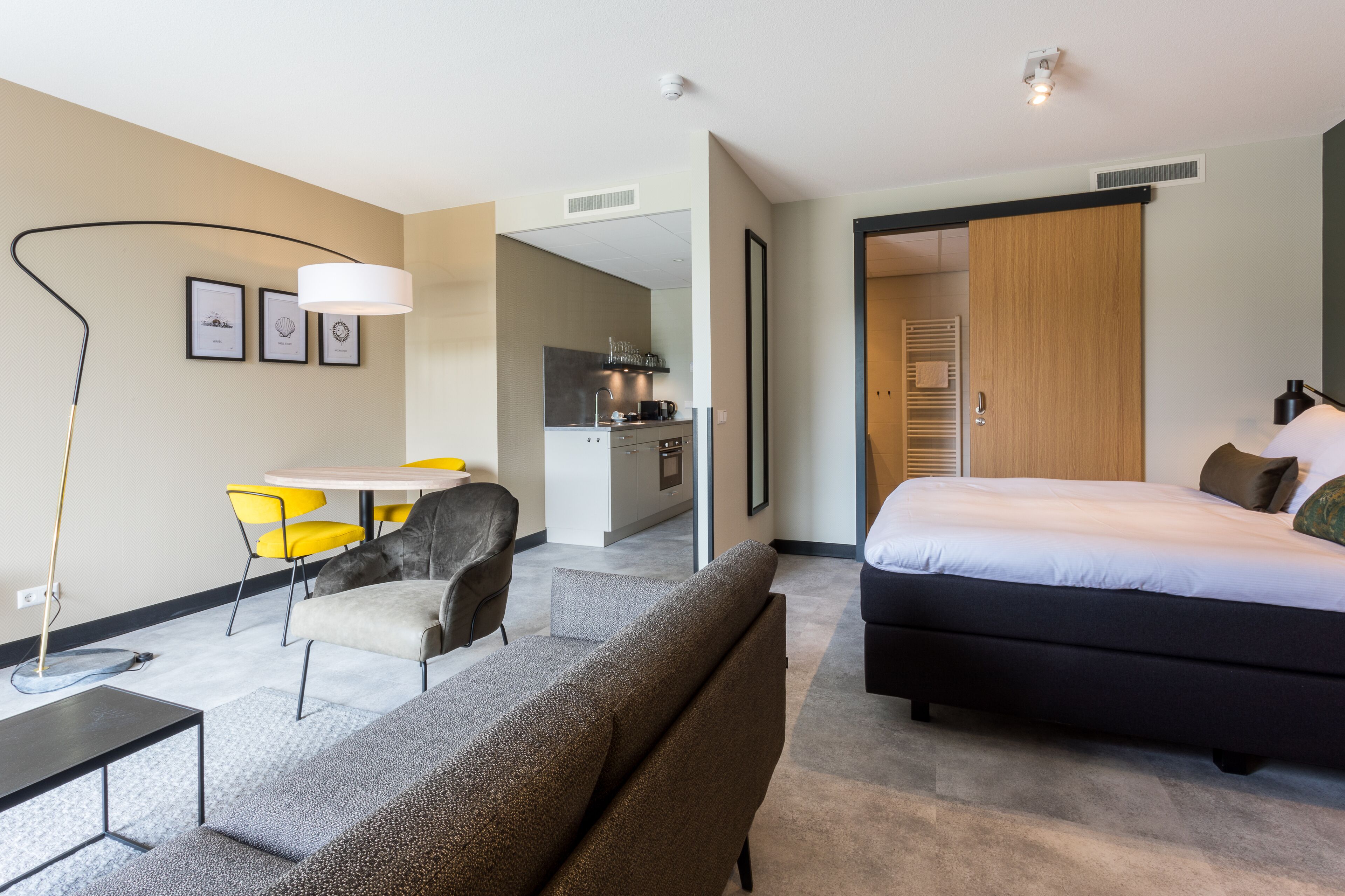 Une chambre d’hôtel moderne avec un grand lit, un canapé, un coin repas avec des chaises jaunes et une kitchenette.