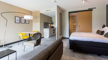Une chambre d’hôtel moderne avec un grand lit, un canapé, un coin repas avec des chaises jaunes et une kitchenette.